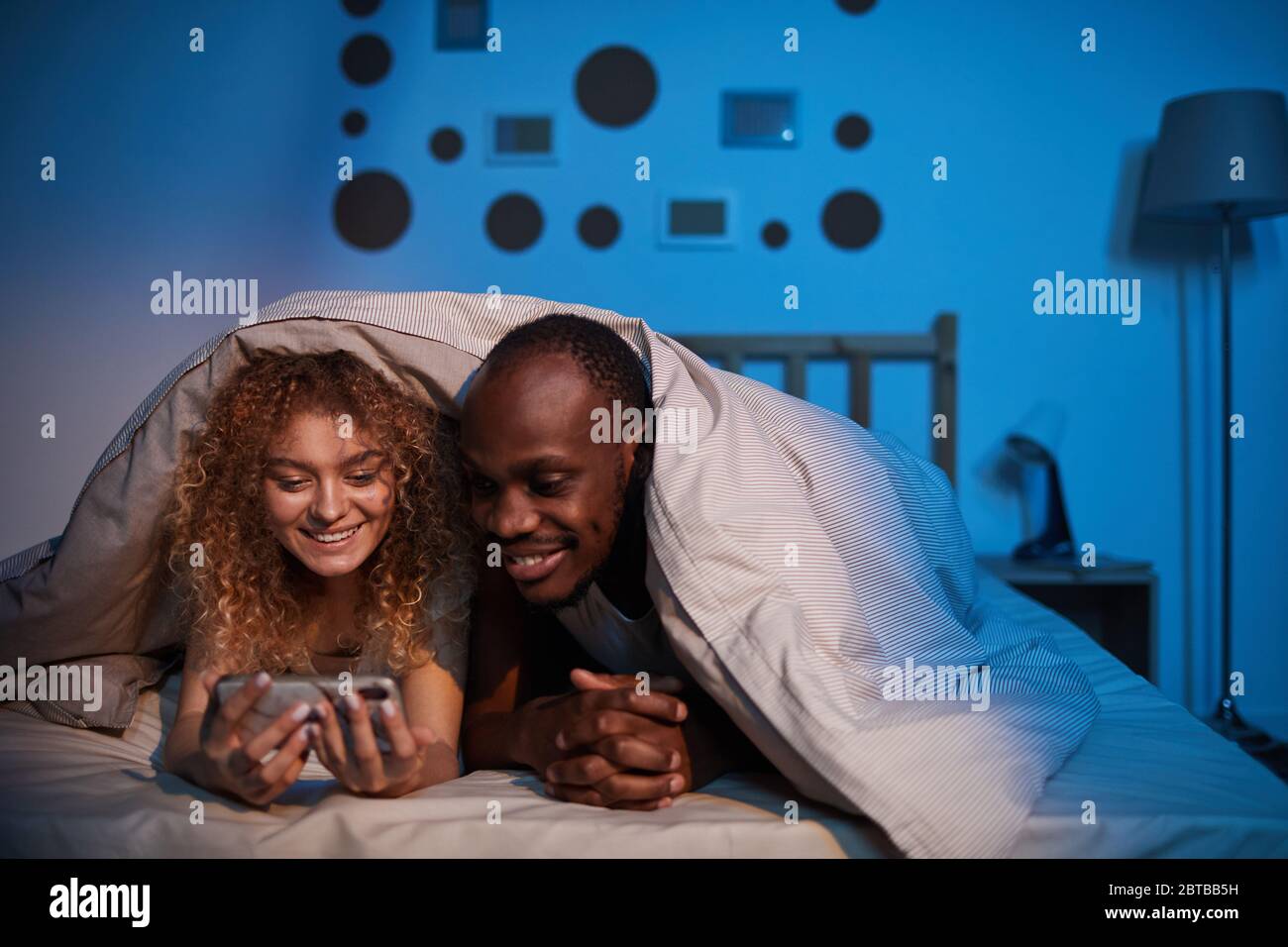 Portrait d'un couple de race mixte sans souci regardant l'écran du smartphone et souriant en étant couché la nuit dans le lit, espace de copie Banque D'Images