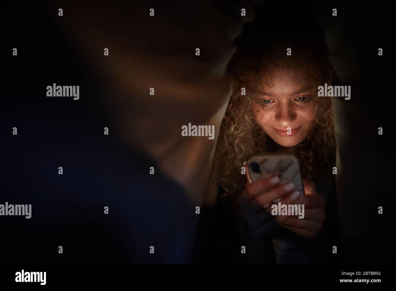 Portrait d'une jeune femme aux cheveux bouclés regardant l'écran du smartphone et souriant en étant allongé dans un lit dans une pièce sombre, espace de copie Banque D'Images