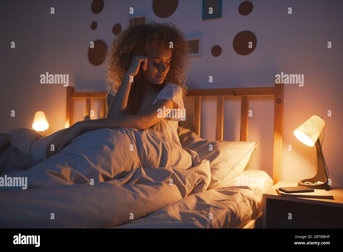 Portrait d'une jeune femme aux cheveux bouclés assise dans un lit confortable la nuit et regardant un smartphone en attendant un appel ou un SMS, espace de copie Banque D'Images