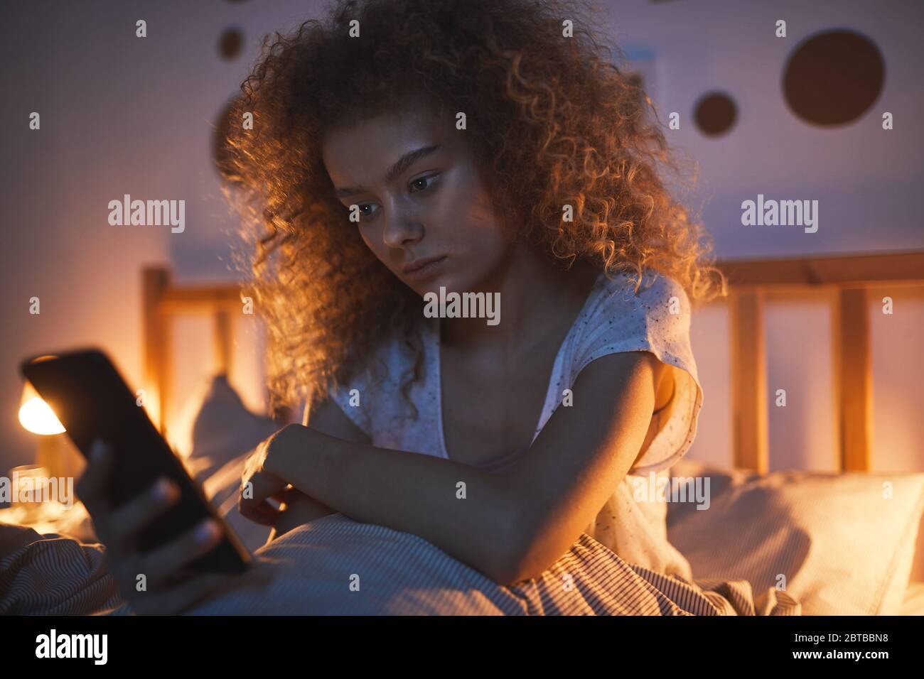Portrait de la jeune femme aux cheveux bouclés assise dans un lit confortable la nuit et tenant un téléphone portable, un espace de copie Banque D'Images