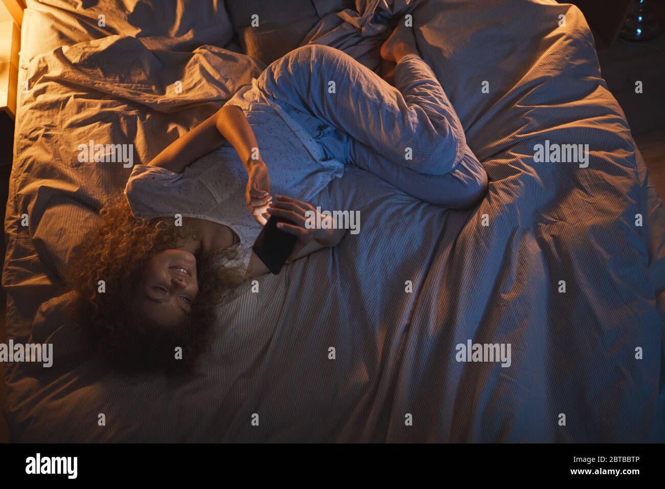 Vue de dessus en bas chez une jeune femme qui s'est allongée sur le lit la nuit et qui utilise un smartphone tout en envoyant un SMS à un ami, espace de copie Banque D'Images