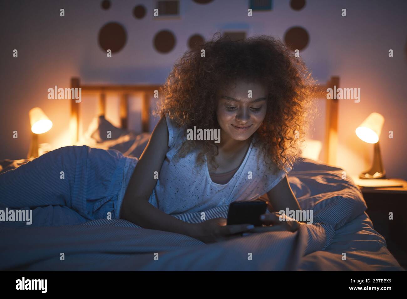 Portrait de la jeune femme aux cheveux bouclés, couchée sur le lit la nuit et souriante avec joie tout en regardant l'écran du smartphone, espace de copie Banque D'Images