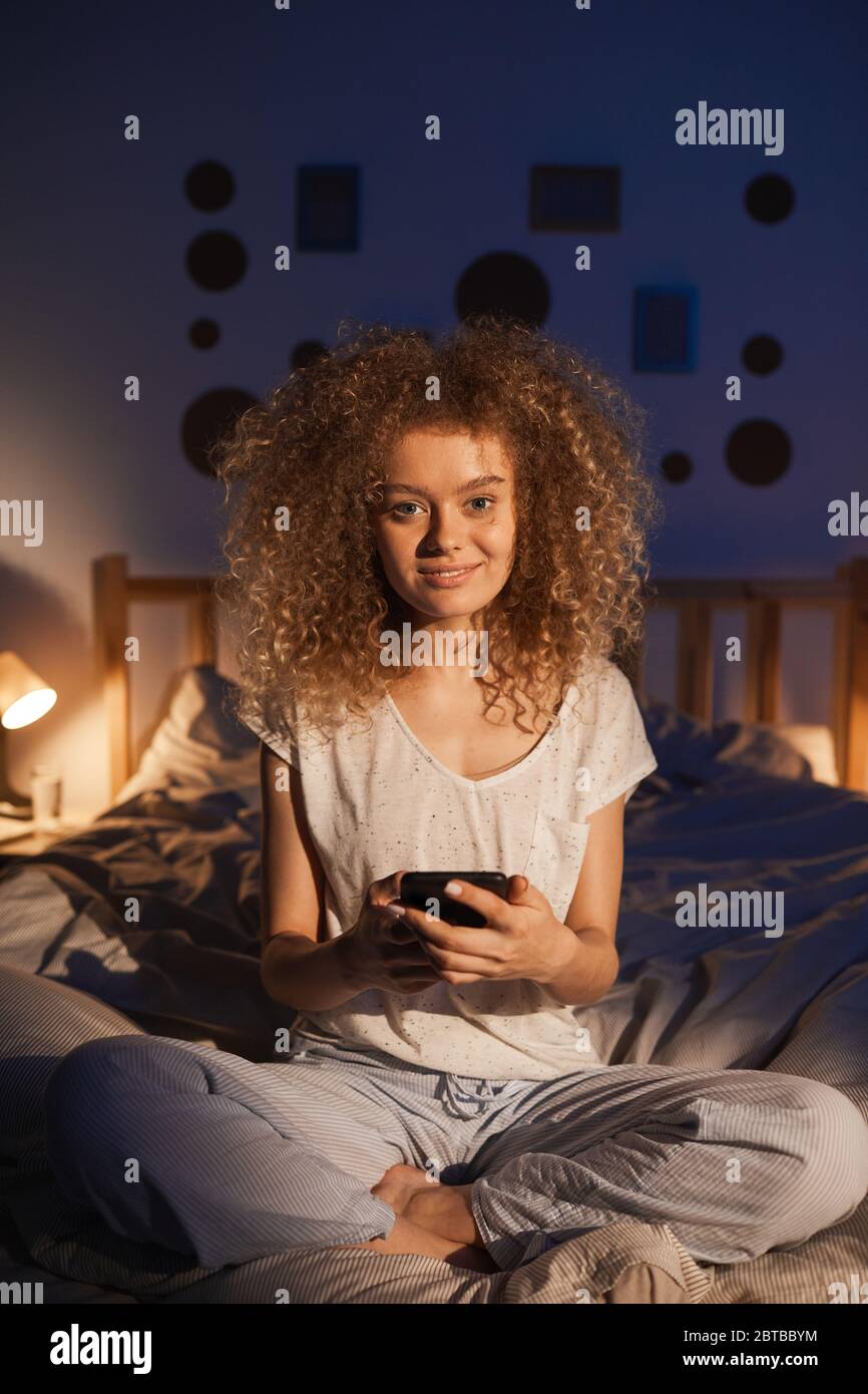 Portrait vertical complet de la jeune femme aux cheveux bouclés assise sur le lit tôt le matin et souriant à l'appareil photo avec joie Banque D'Images