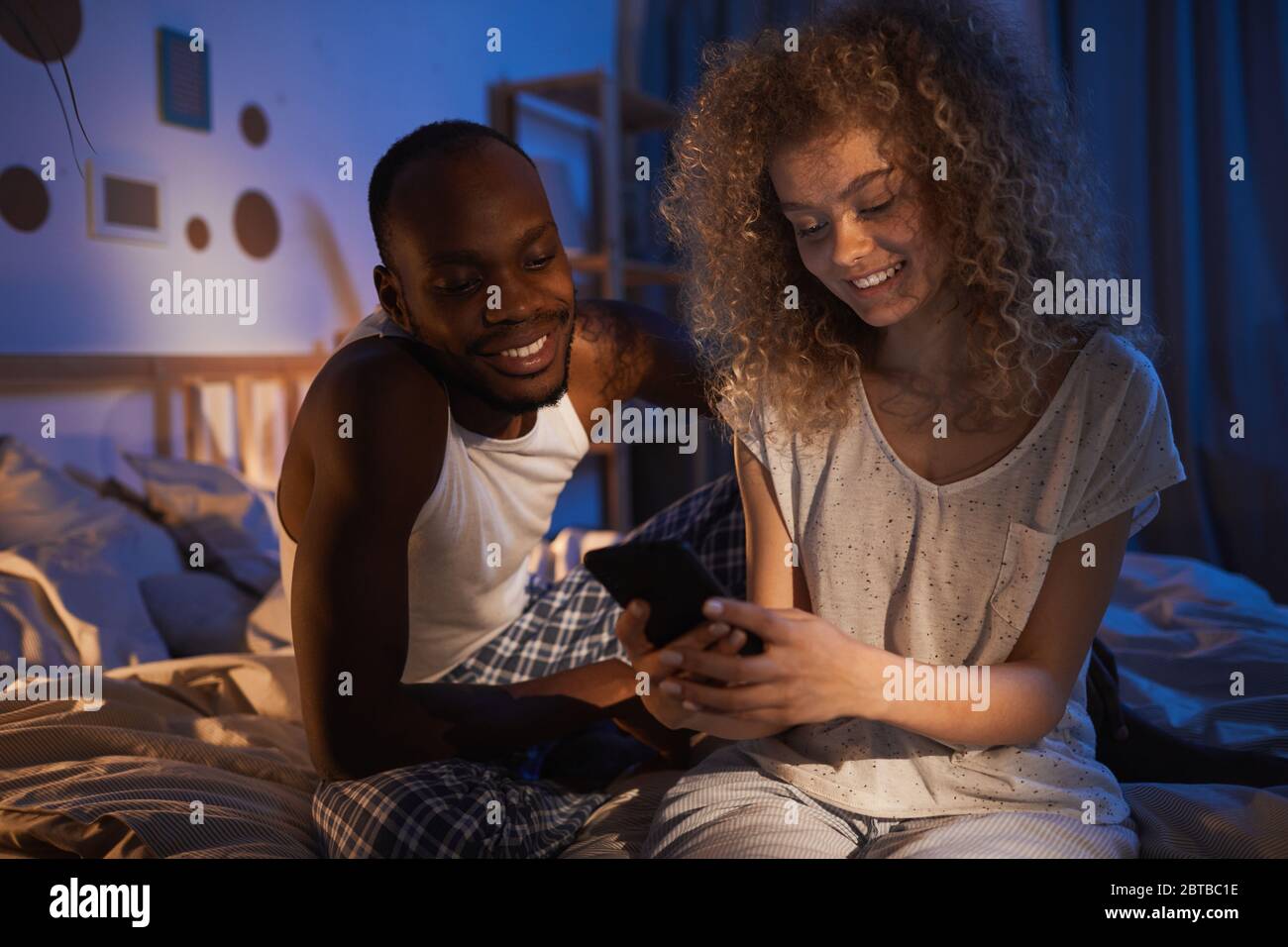 Portrait d'un jeune couple de course mixte assis sur le lit la nuit et souriant avec joie tout en regardant l'écran du smartphone Banque D'Images