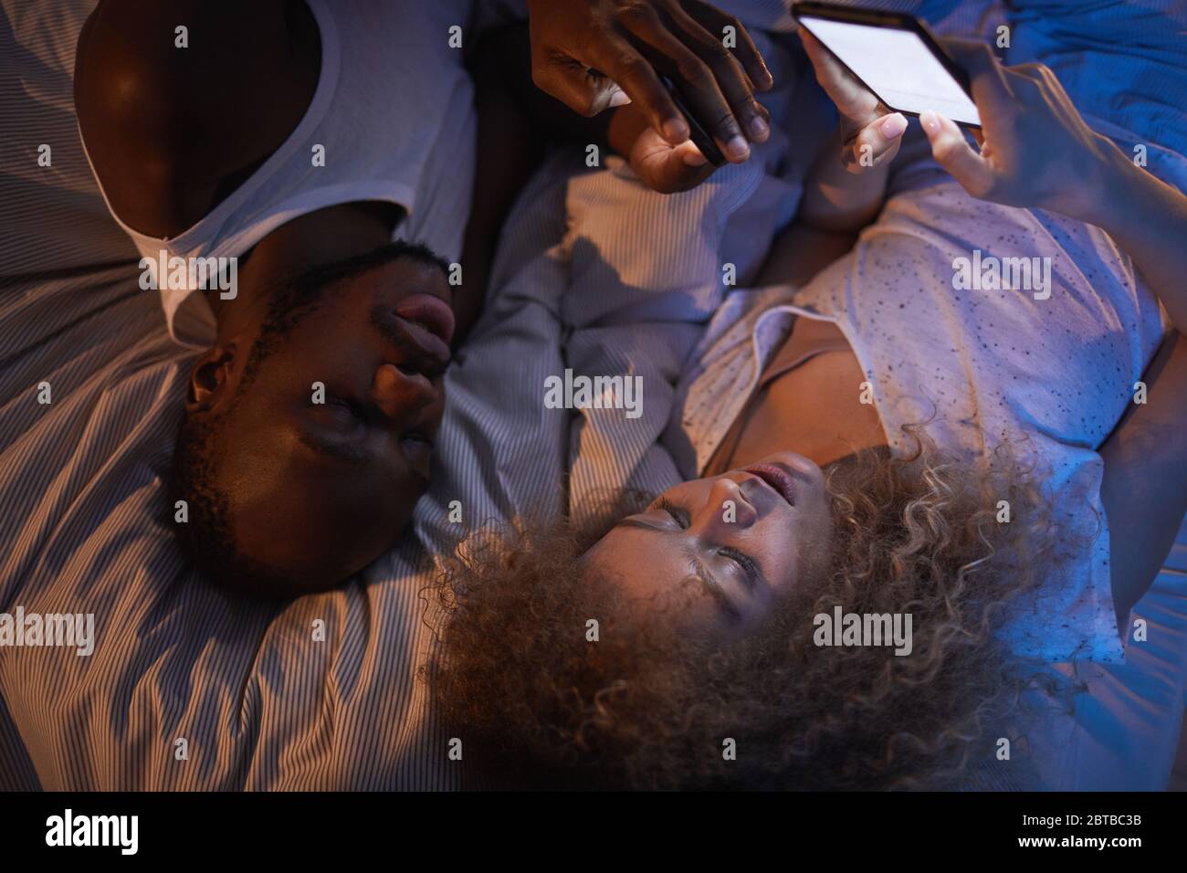 Portrait grand angle de jeunes couples de course mixte allongé sur le lit la nuit en regardant les écrans de smartphone pendant les conversations Banque D'Images