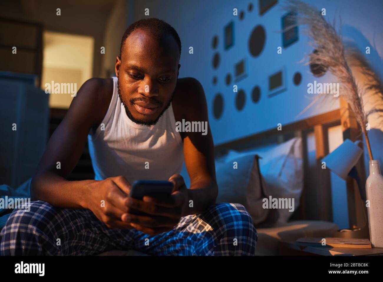 Portrait à angle bas d'un homme afro-américain adulte utilisant un smartphone assis sur le lit la nuit, espace de copie Banque D'Images