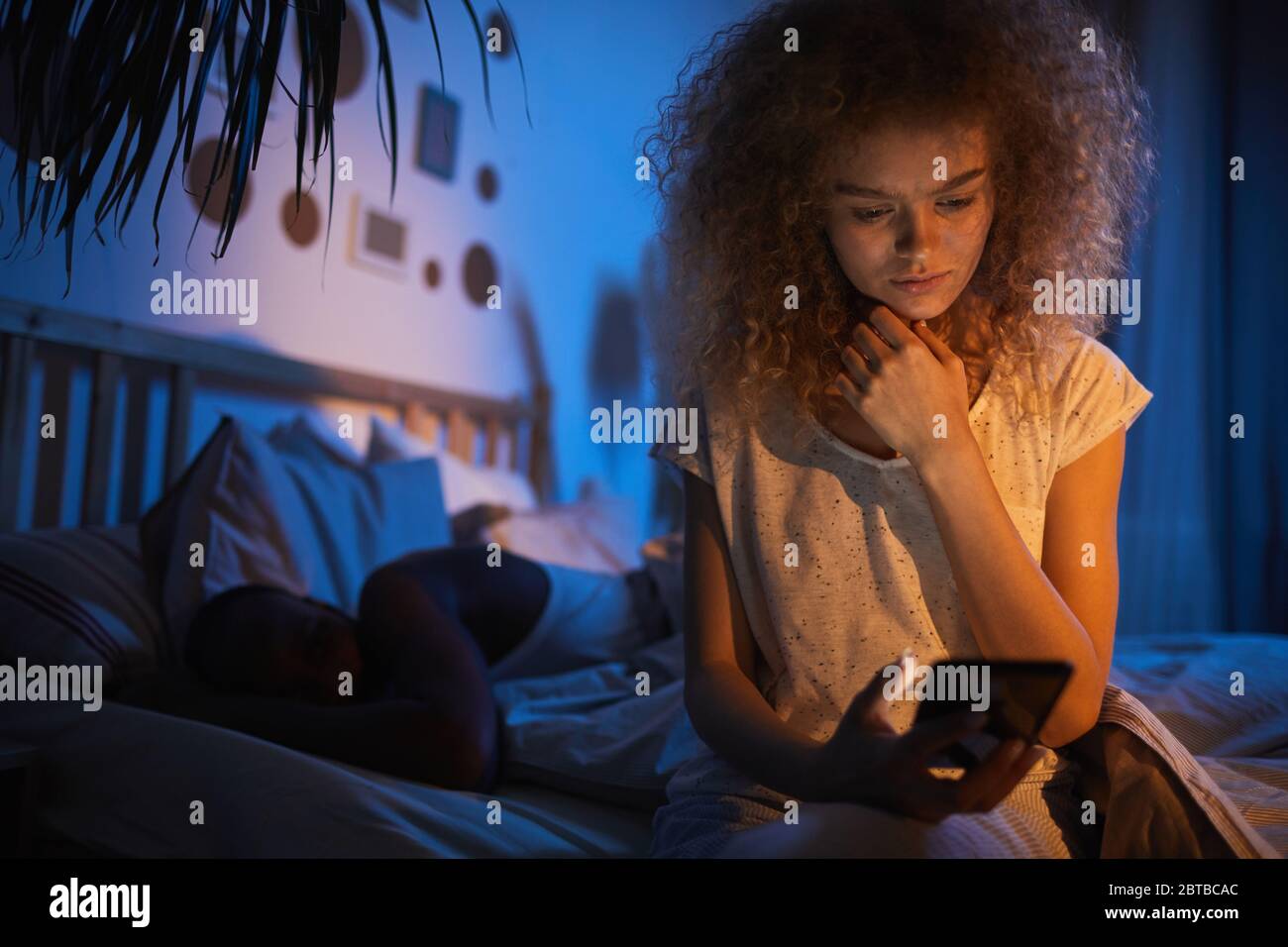 Portrait de la jeune femme aux cheveux bouclés assise sur un lit familial la nuit et tenant un smartphone, en regardant nerveux, espace de copie Banque D'Images