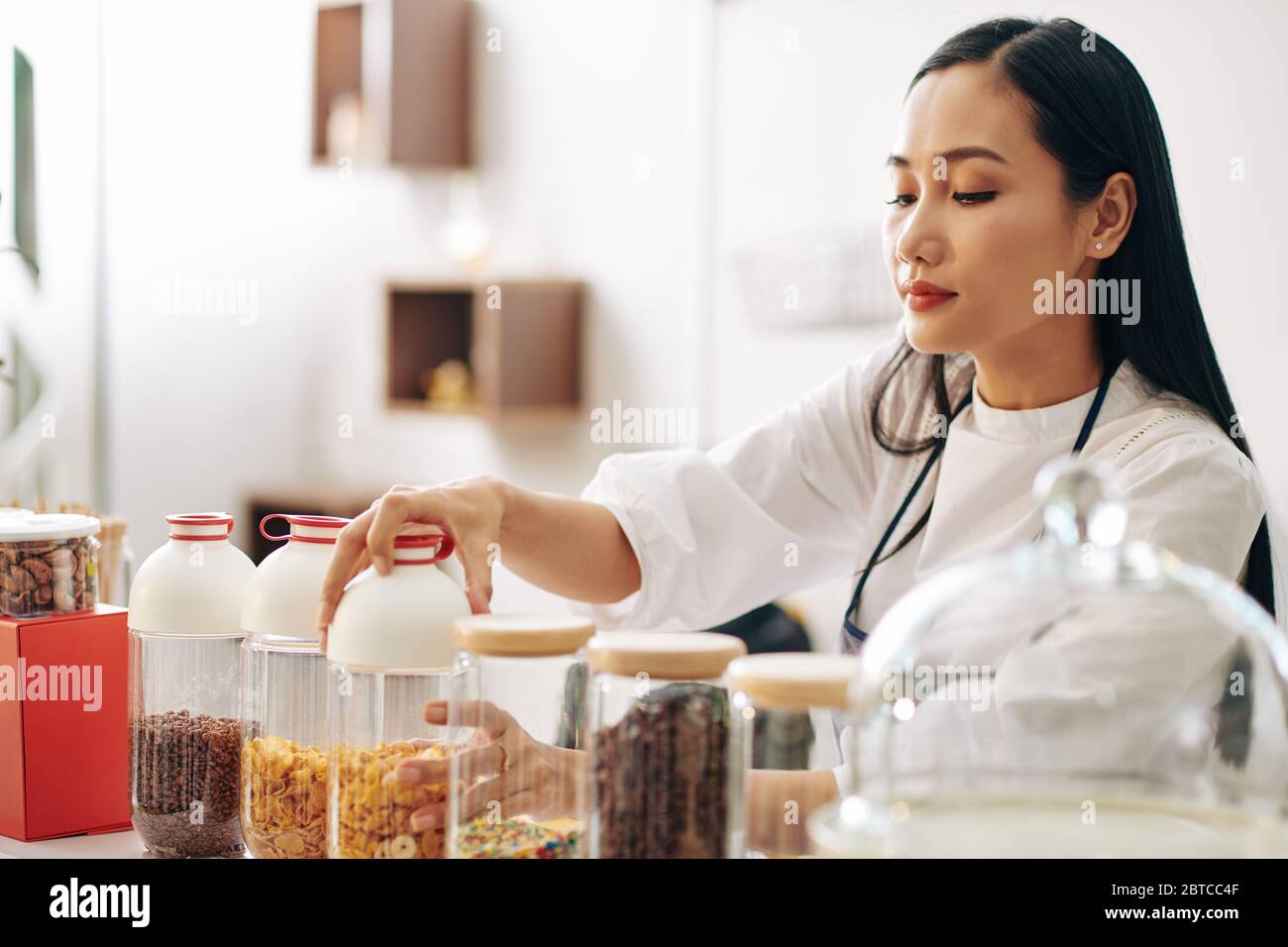 Joli jeune serveuse ouvrant le contenant avec des céréales lors de la préparation du petit déjeuner pour le client Banque D'Images