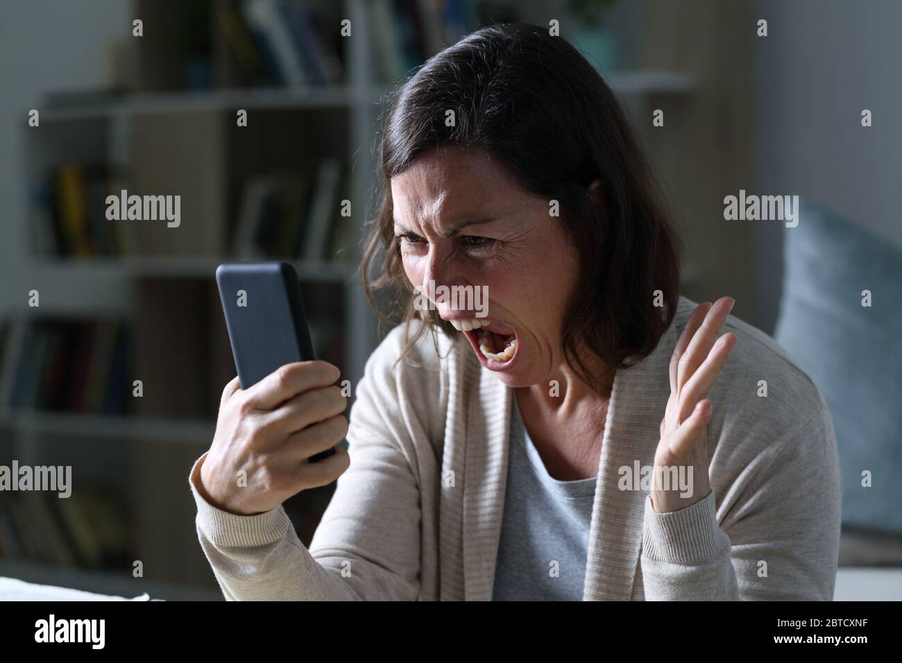 Une femme adulte en colère criant en regardant un smartphone assis dans le salon la nuit à la maison Banque D'Images