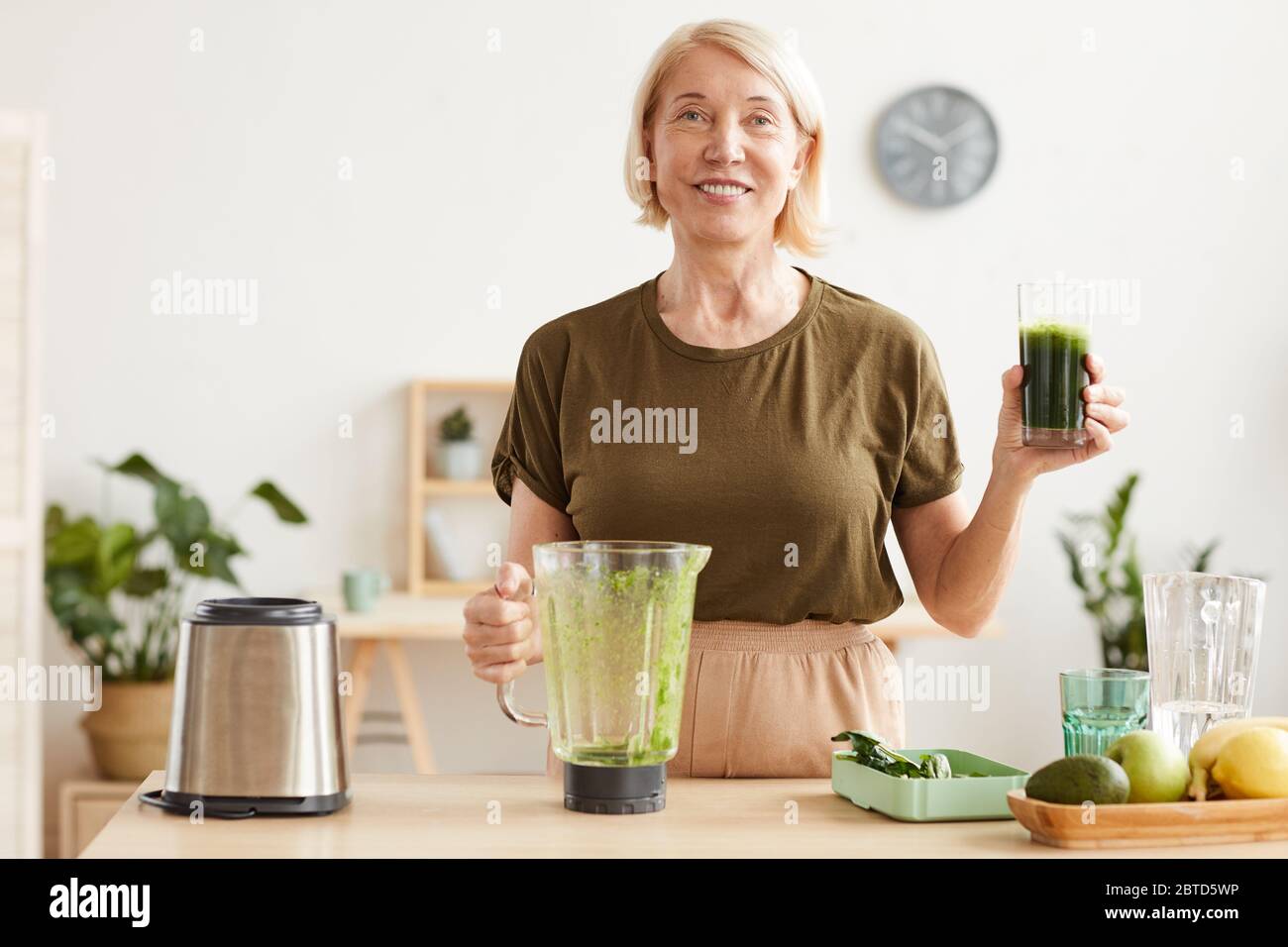 Portrait d'une femme mûre aux cheveux blonds souriant à l'appareil photo et buvant du jus de légumes frais tout en étant debout dans la cuisine Banque D'Images