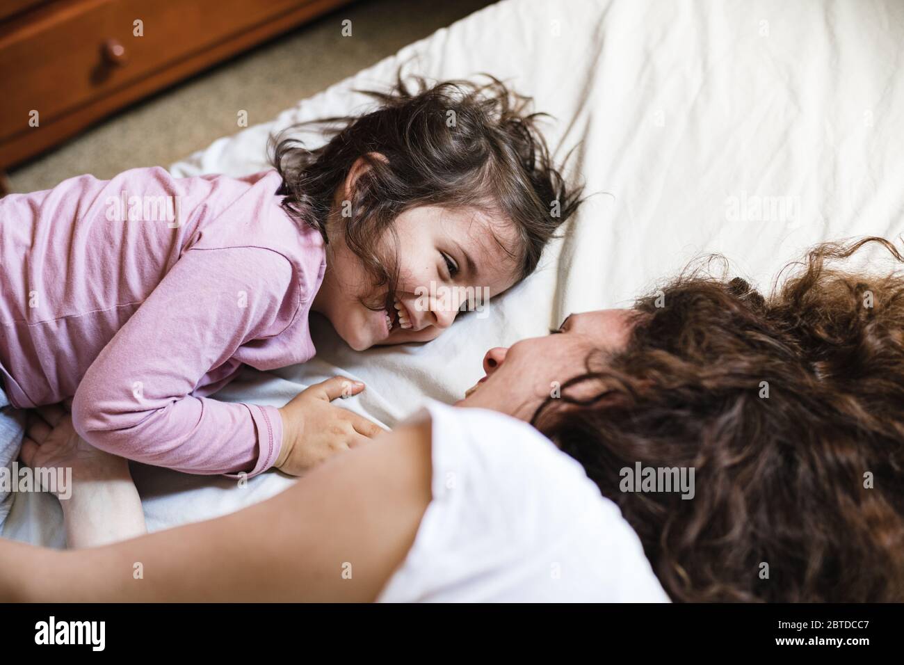 Une petite fille qui s'est allongée dans le lit de sa mère rit pendant qu'elle a un grand temps à jouer autour Banque D'Images