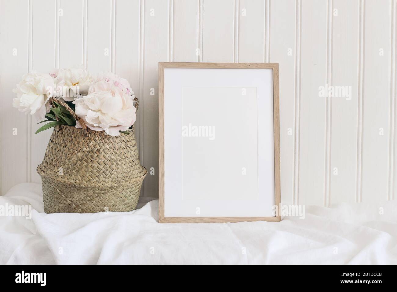 Maquette de cadre en bois beige vierge. Table artistique encore la composition de vie avec des fleurs de pivoine rose dans le panier de paille. Fond de mur blanc en bois, Banque D'Images