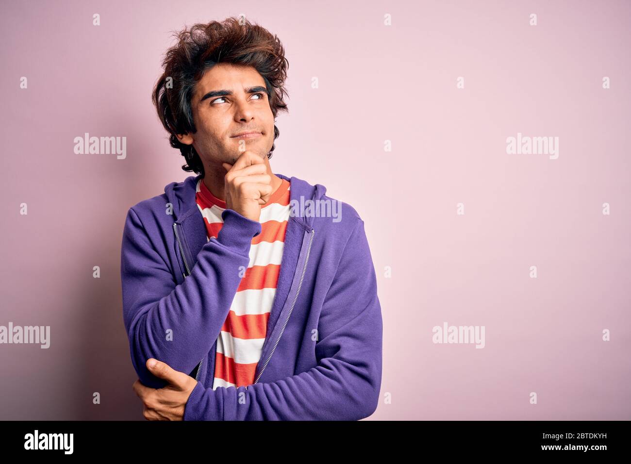 Jeune homme portant un t-shirt rayé et un sweat-shirt violet sur fond rose isolé avec la main sur le menton pensant à la question, l'expression pensive. SM Banque D'Images