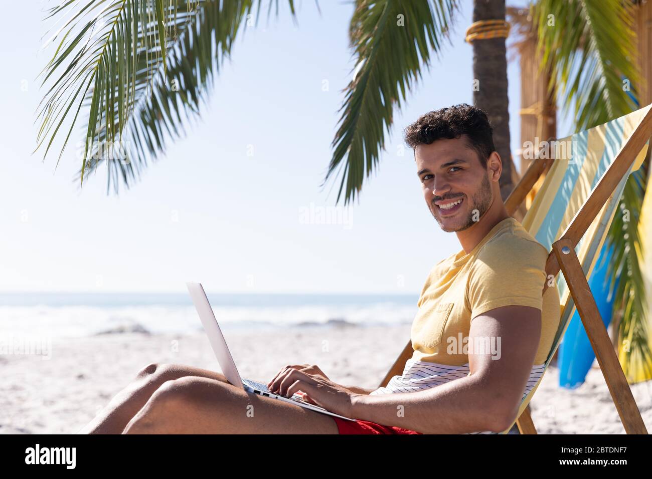 Homme caucasien assis sur une chaise longue et utilisant un ordinateur portable à la plage Banque D'Images