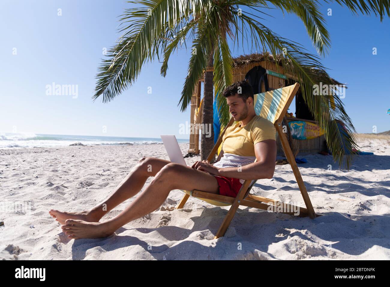 Homme caucasien assis sur une chaise longue et utilisant un ordinateur portable à la plage Banque D'Images