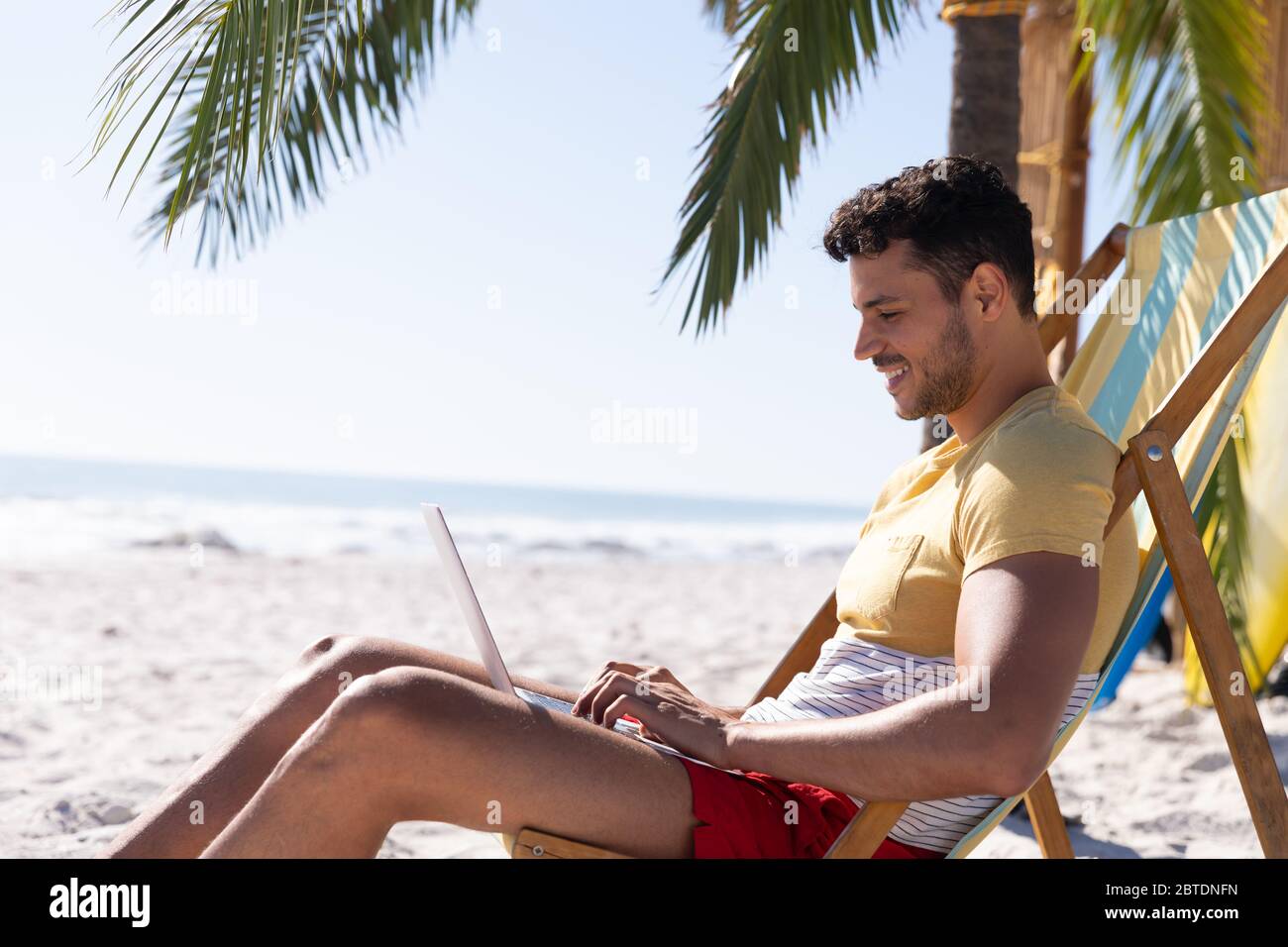 Homme caucasien assis sur une chaise longue et utilisant un ordinateur portable à la plage Banque D'Images