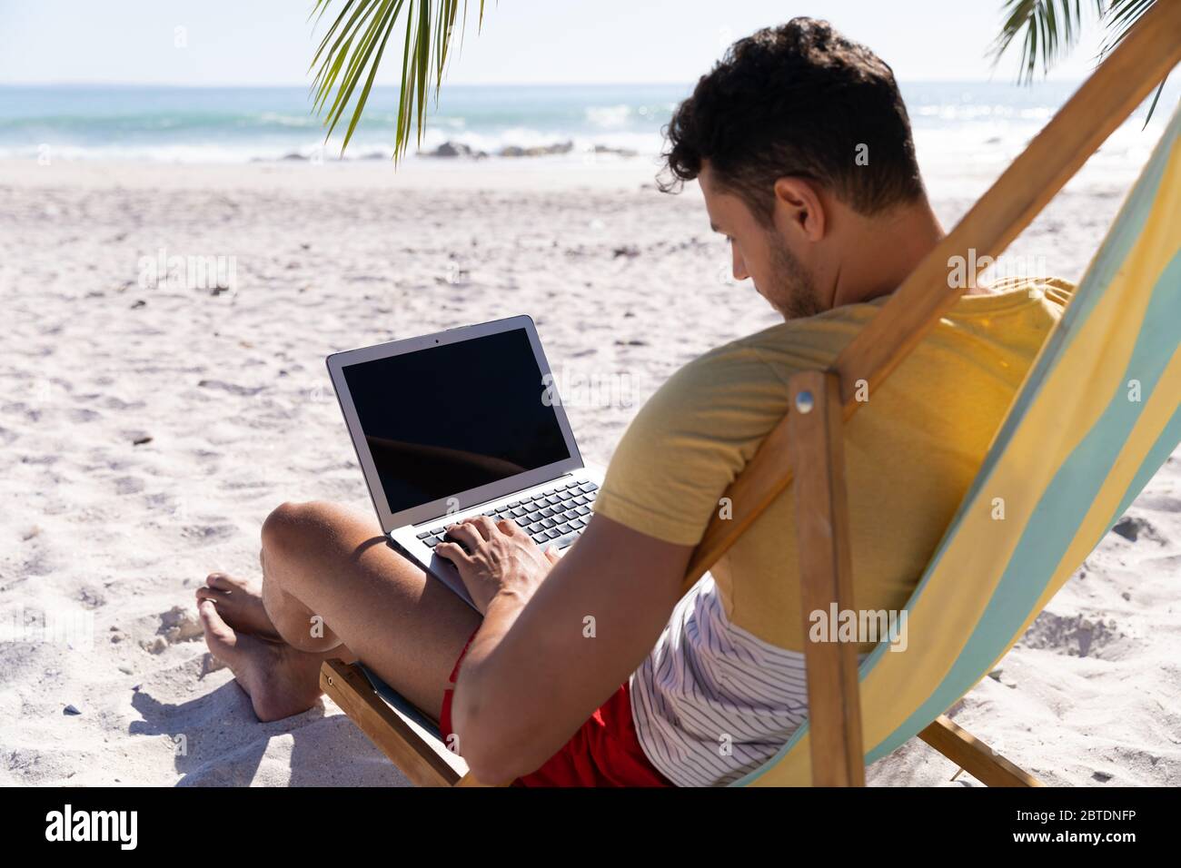 Homme caucasien assis sur une chaise longue et utilisant un ordinateur portable à la plage Banque D'Images