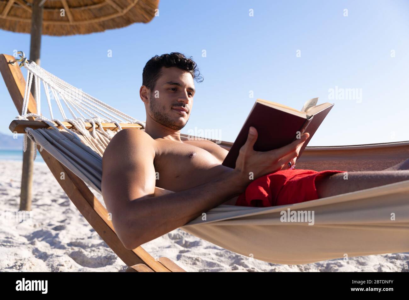 Homme caucasien allongé sur un hamac et lisant un livre à la plage. Banque D'Images