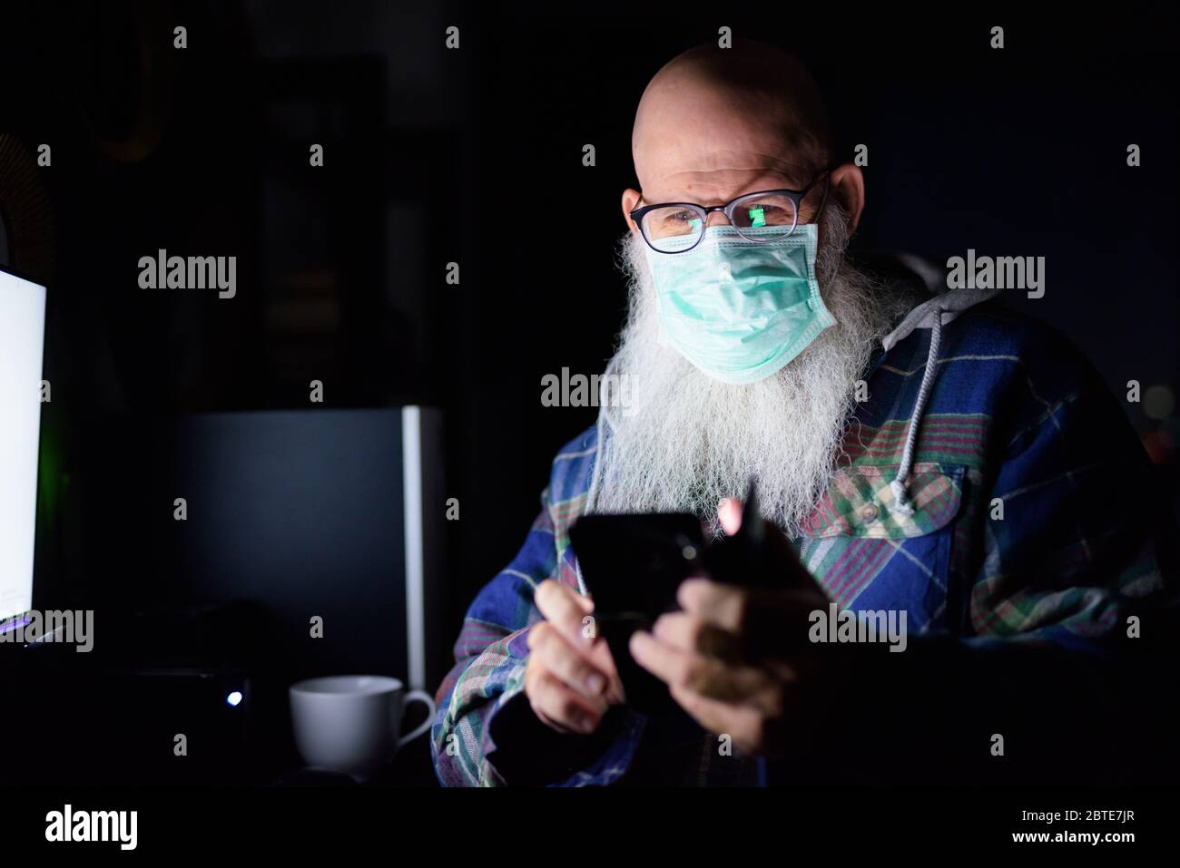 Visage d'un homme à barbe chauve mature avec masque en utilisant le  téléphone tout en travaillant de la maison tard dans la nuit Photo Stock -  Alamy