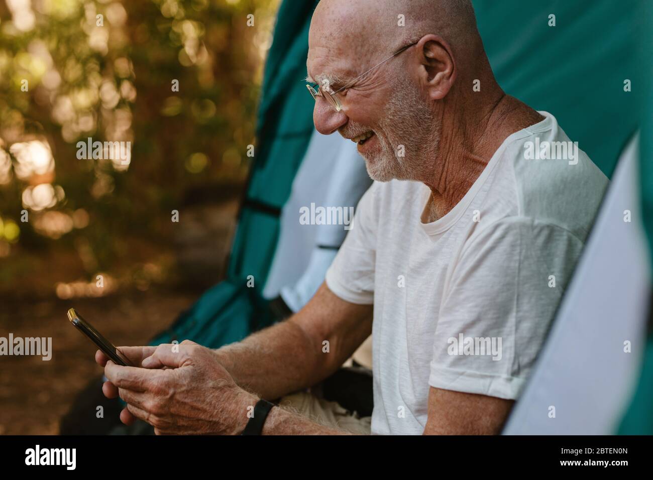 Homme senior souriant assis dans une tente à l'aide de son téléphone portable. Homme à la retraite campant dans la nature lisant un message texte sur son téléphone cellulaire. Banque D'Images