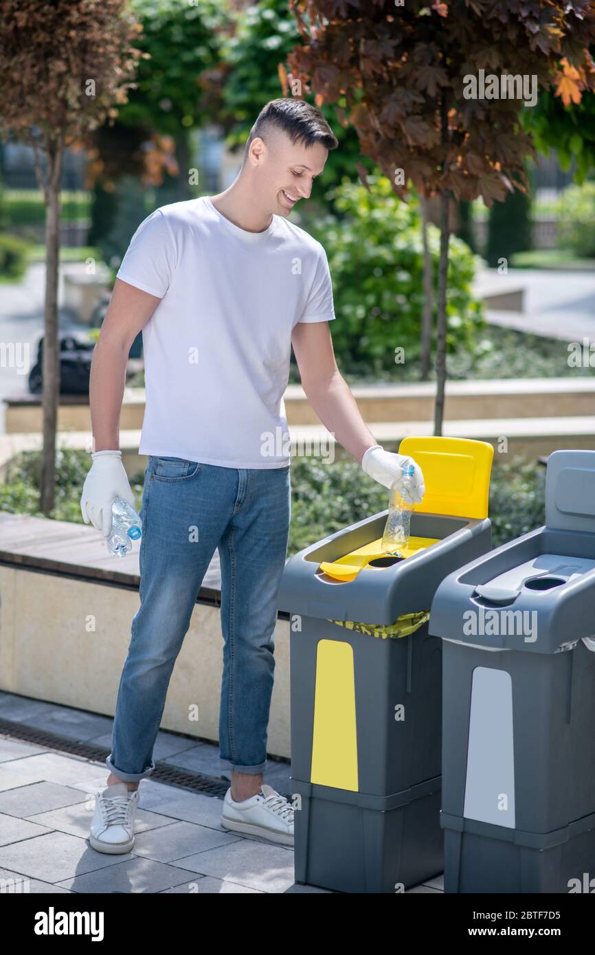 Homme à poil sombre en gants de protection, jetant des bouteilles en plastique ouvertes dans un conteneur de déchets Banque D'Images