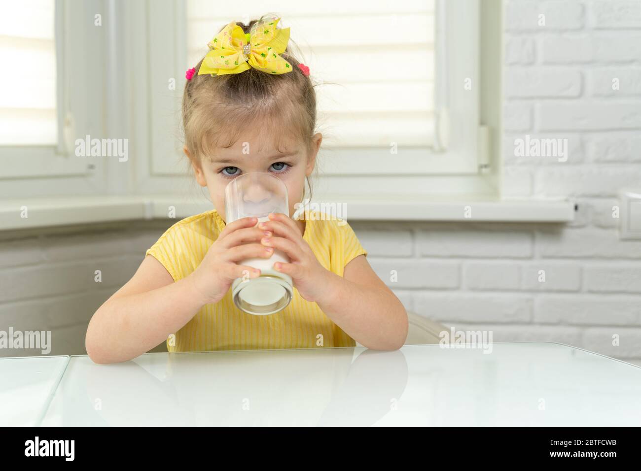 une petite fille dans un chemisier jaune boit du lait dans un verre dans la cuisine Banque D'Images