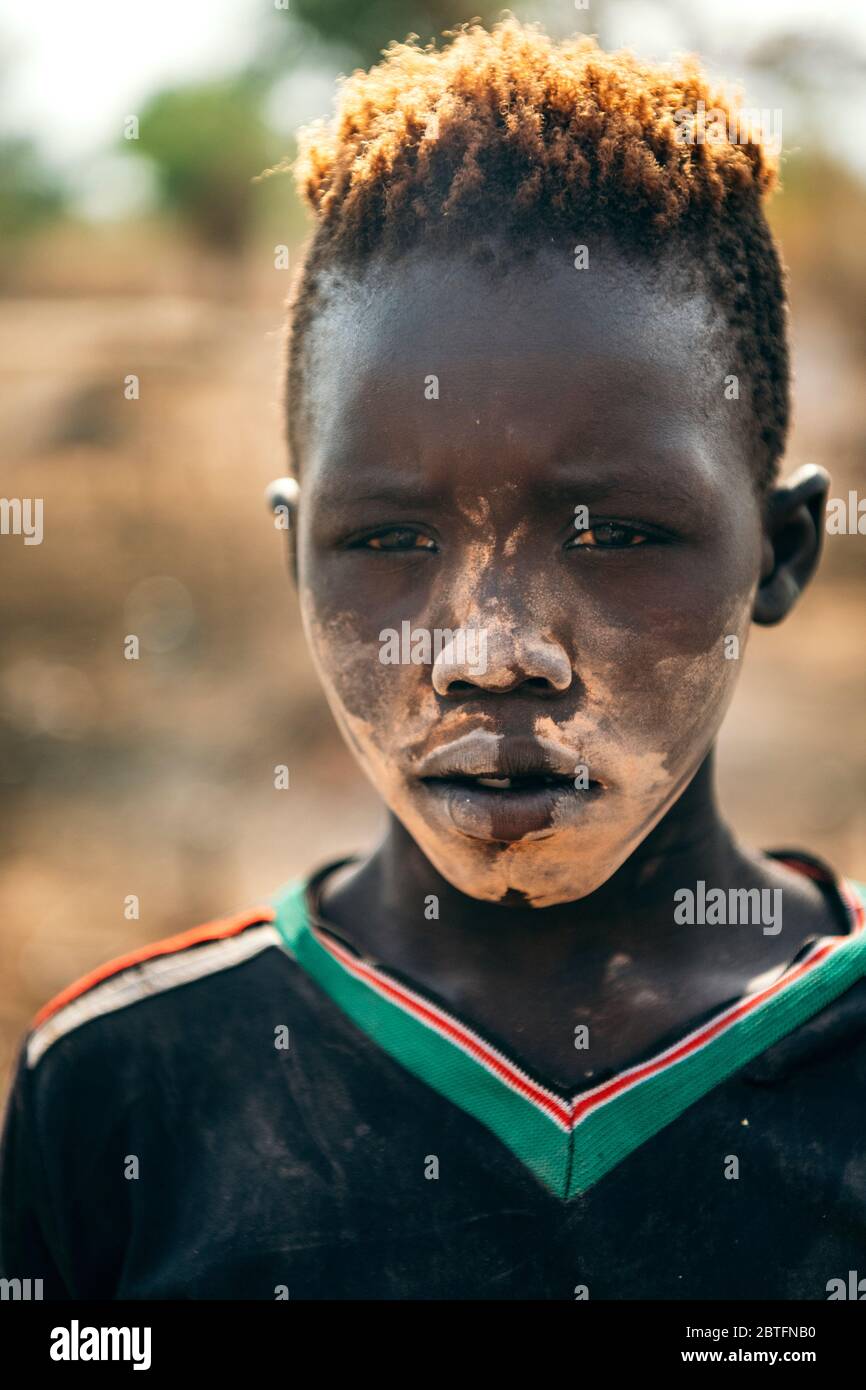 TRIBU MUNDARI, SOUDAN DU SUD - 11 MARS 2020 : jeune garçon avec la saleté rituelle sur le visage et dans le t-shirt regardant la caméra tout en vivant dans le village de la tribu Mundari Banque D'Images