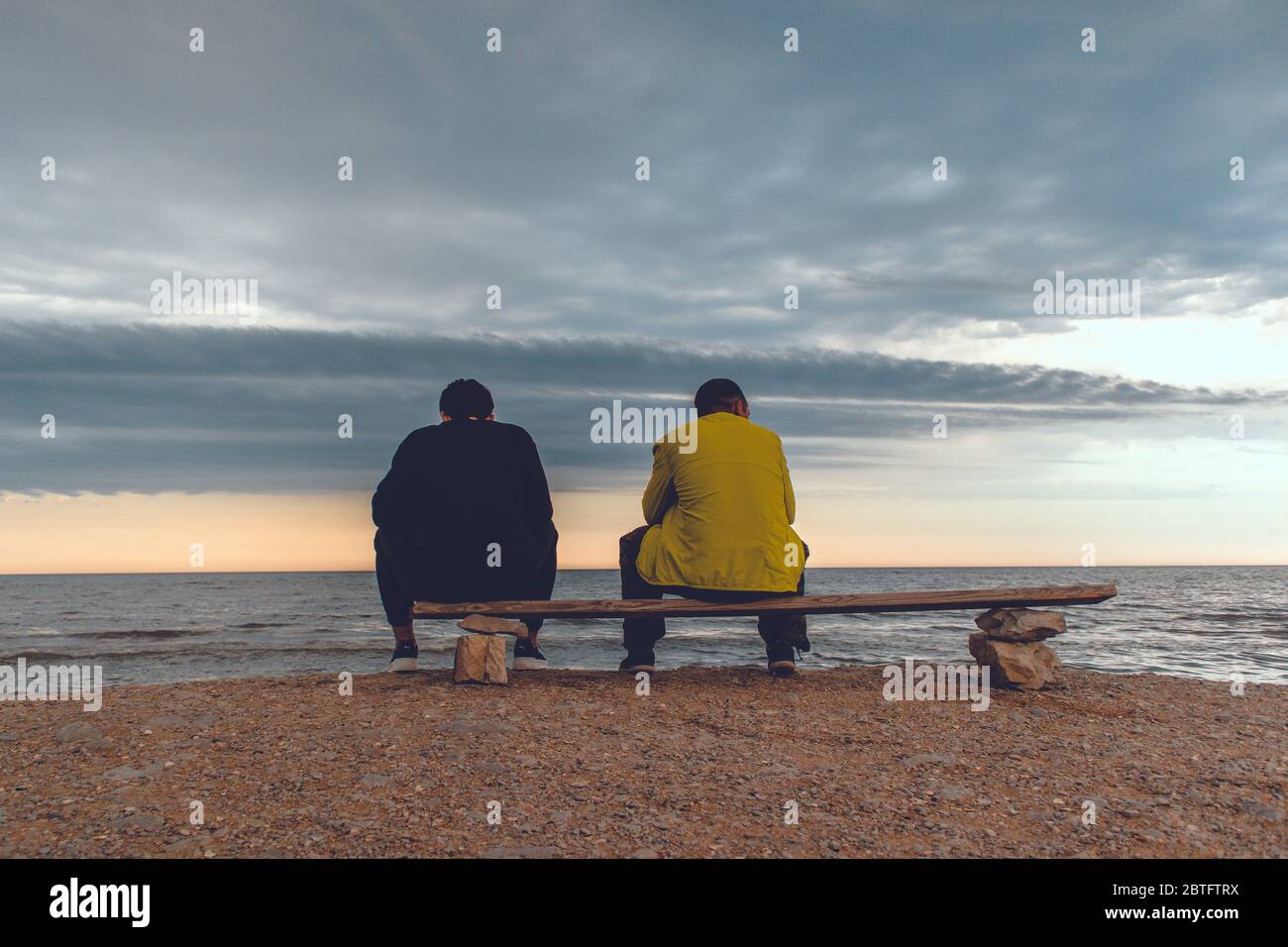 Deux hommes s'assoient sur un banc près d'une plage et regardent l'océan avec des nuages épiques. Banque D'Images