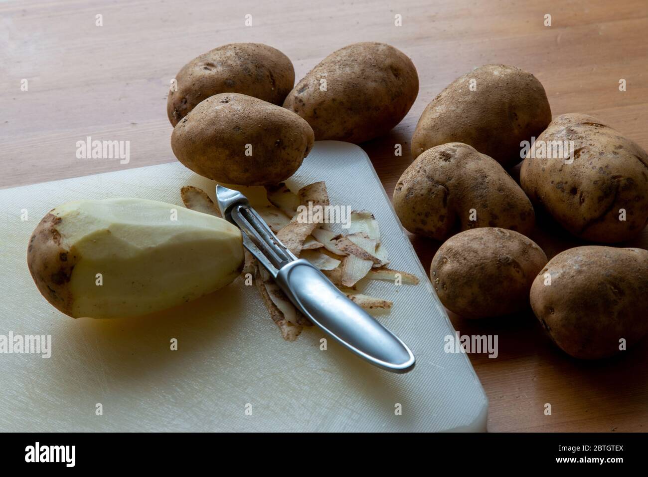 Une pomme de terre brute est pelée sur une planche à découper avec un outil de pelage en métal, parmi beaucoup d'autres encore non pelées. Banque D'Images