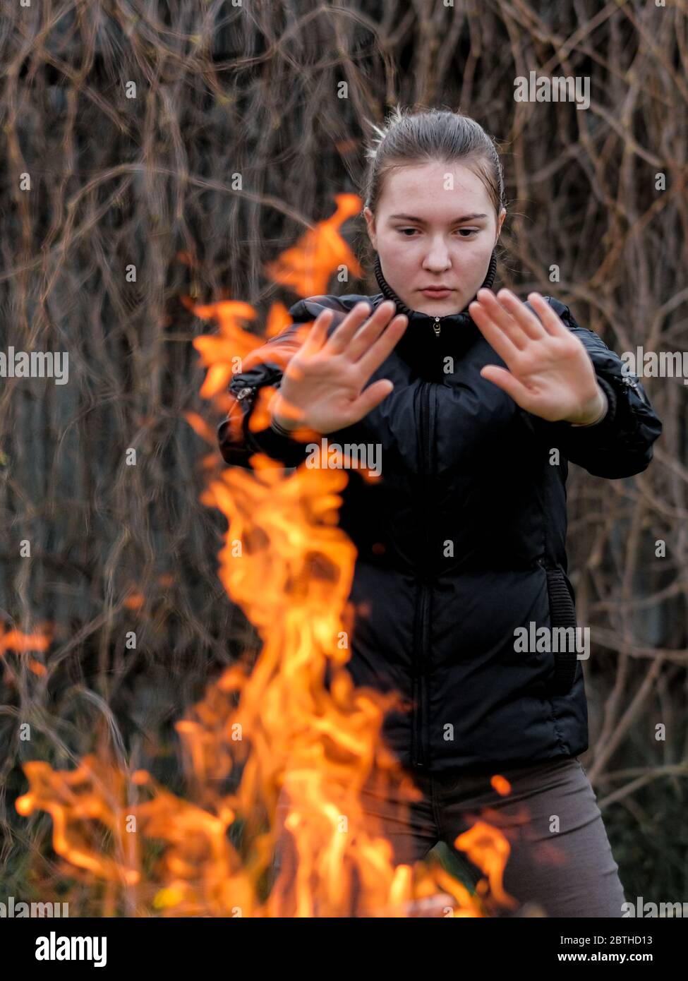 Une jeune fille dans une veste noire qui s'étire les mains réchauffe les paumes par le feu. Flammes orange vif. Arrière-plan flou des branches sèches. Banque D'Images