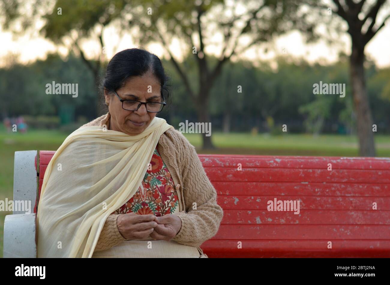 Photo d'une femme indienne âgée assise dans un parc sur un banc rouge qui peeling des arachides et les mangeant. Concept - la vie à la retraite Banque D'Images