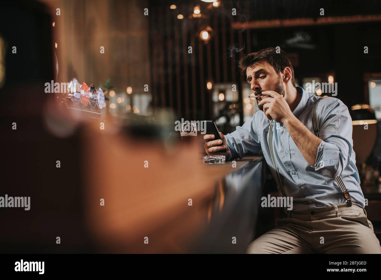 Un jeune homme boit de la bière, fume de la cigarette et utilise le téléphone portable au pub Banque D'Images