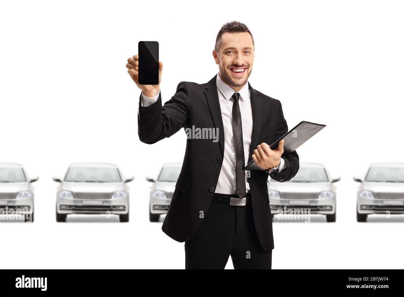 Homme en costume tenant un téléphone mobile et posant devant une rangée de voitures dans une salle d'exposition isolée sur fond blanc Banque D'Images