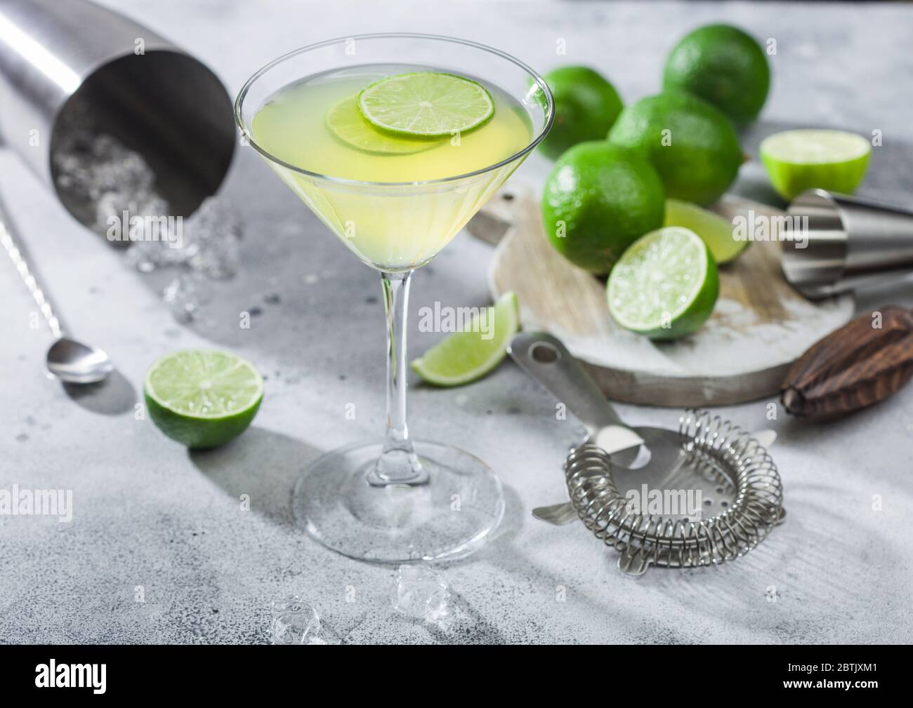 Cocktail de gimlet Kamikaze en verre martini avec tranche de lime et glace sur fond clair avec des limettes fraîches et passoire avec shaker. Cuillère à queue de coktail et gabarit Banque D'Images