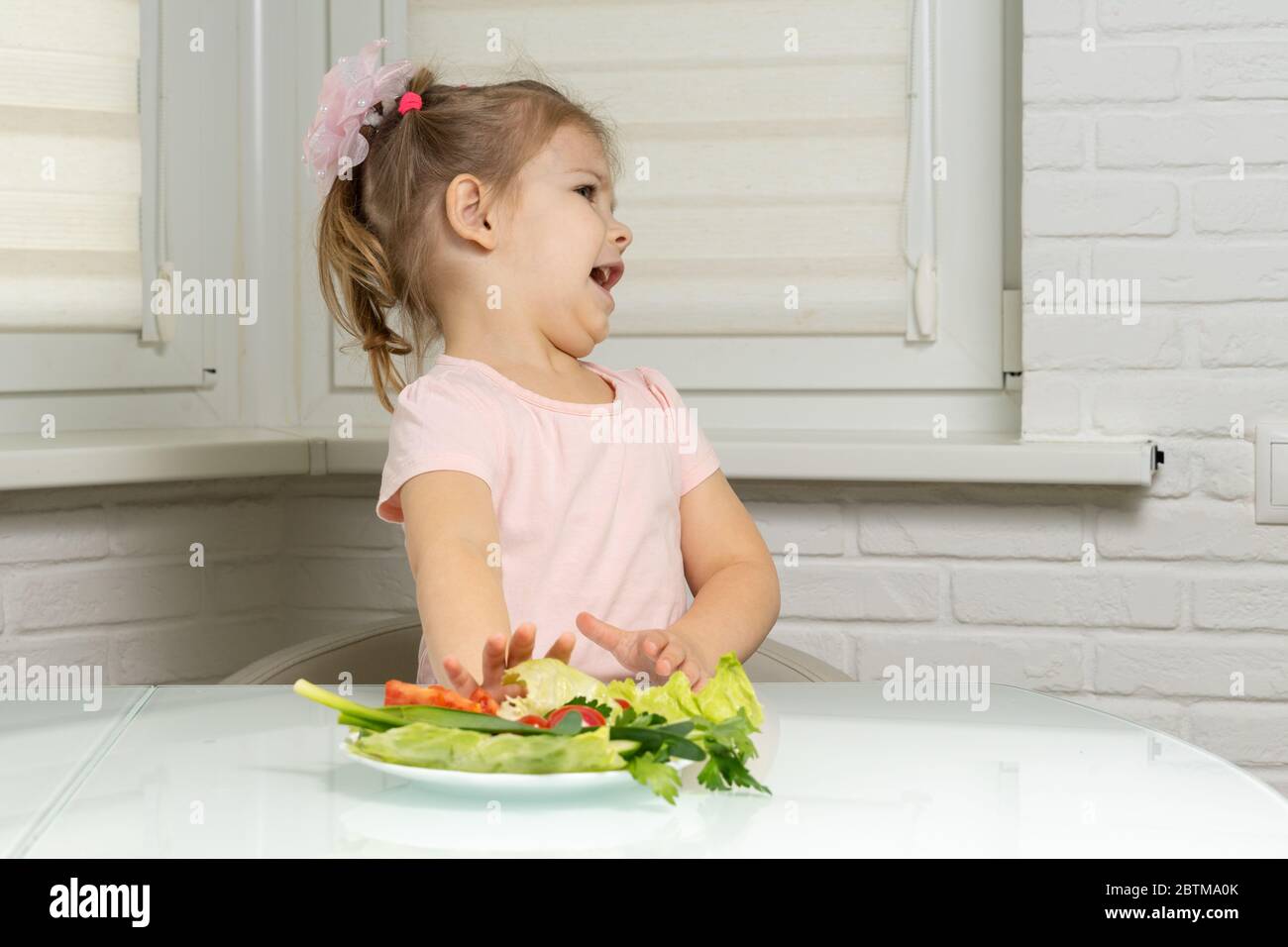 une petite fille s'assoit à une table dans la cuisine et repousse une assiette de légumes. les enfants ne veulent pas manger de légumes Banque D'Images