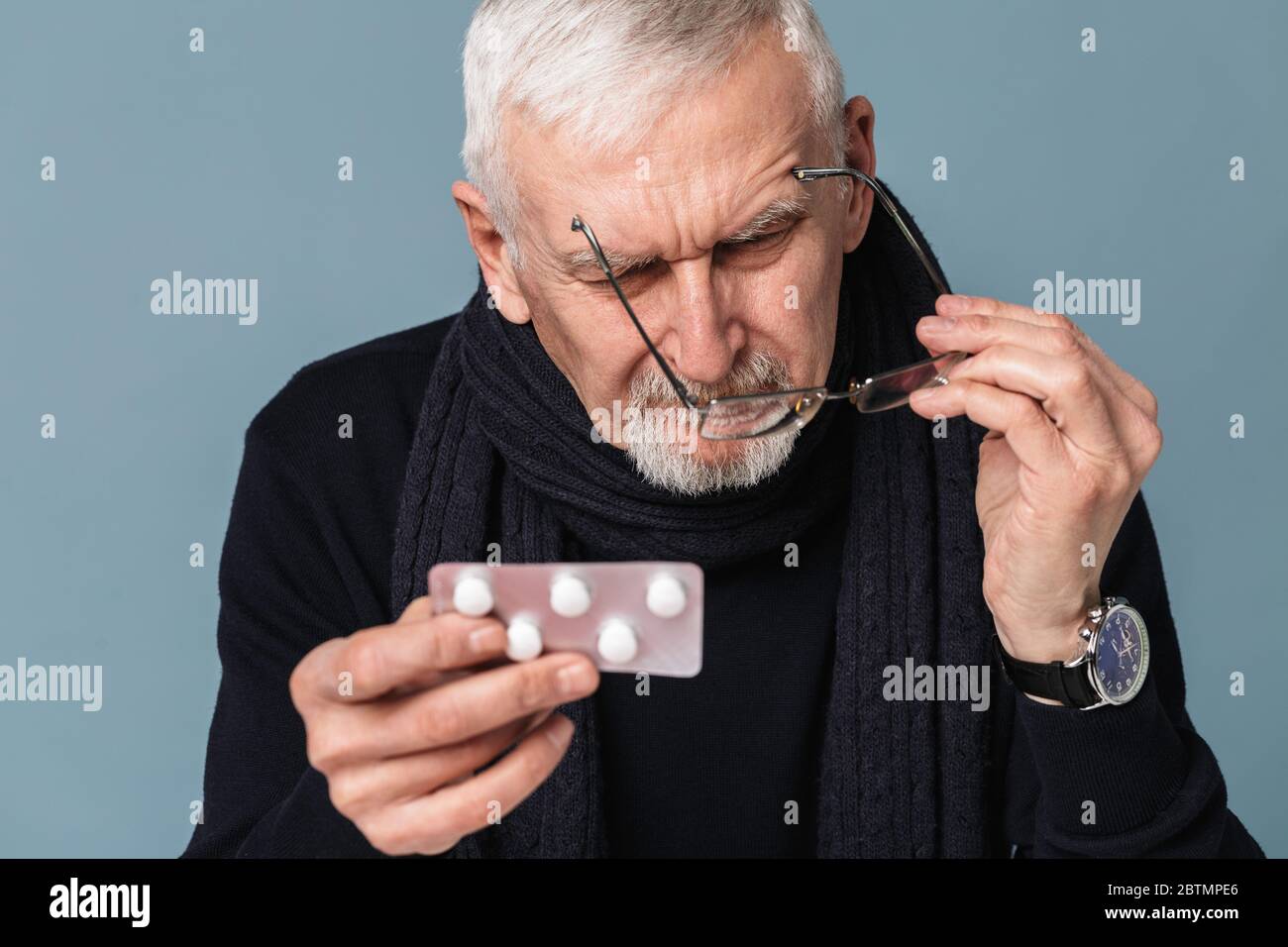 Homme malade pensif vieux avec cheveux gris et barbe dans les lunettes et foulard regardant soigneusement sur les pilules sur fond bleu isolé Banque D'Images