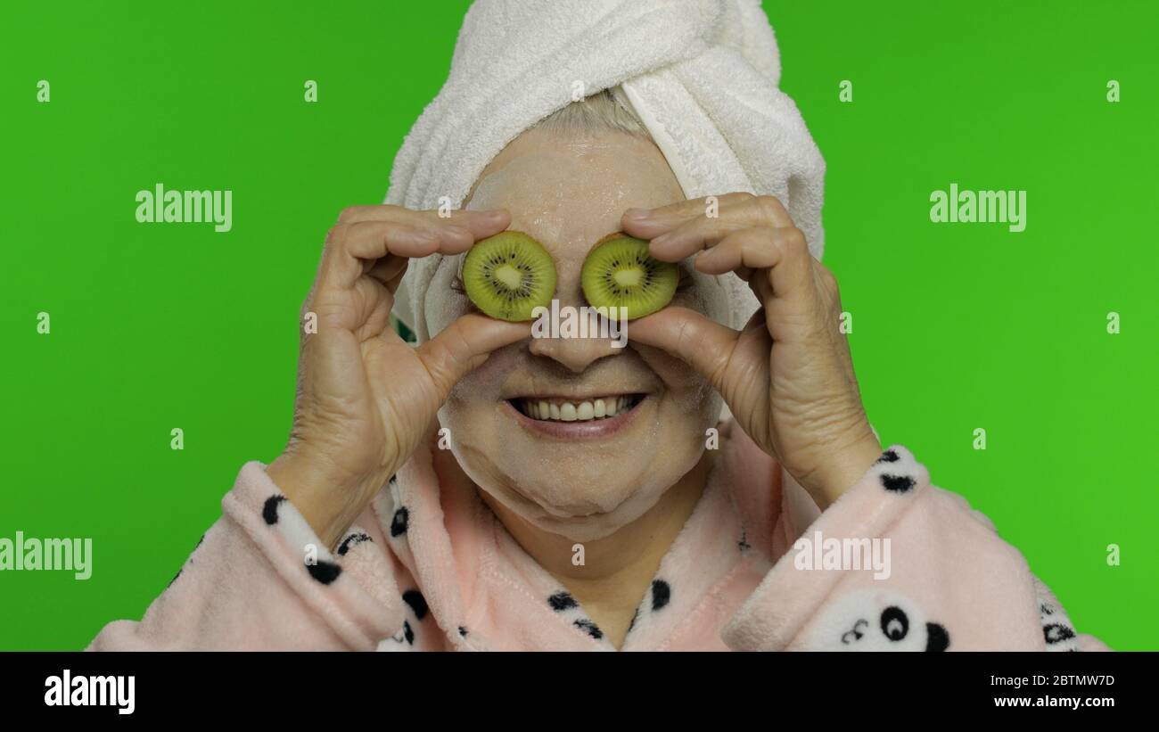 Femme caucasienne adulte sénior grand-mère dans un peignoir et un masque à bulles avec deux moitiés de kiwi appliquées aux yeux isolés sur fond de chroma clé. Procédures de beauté pour adultes grands-parents personnes Banque D'Images