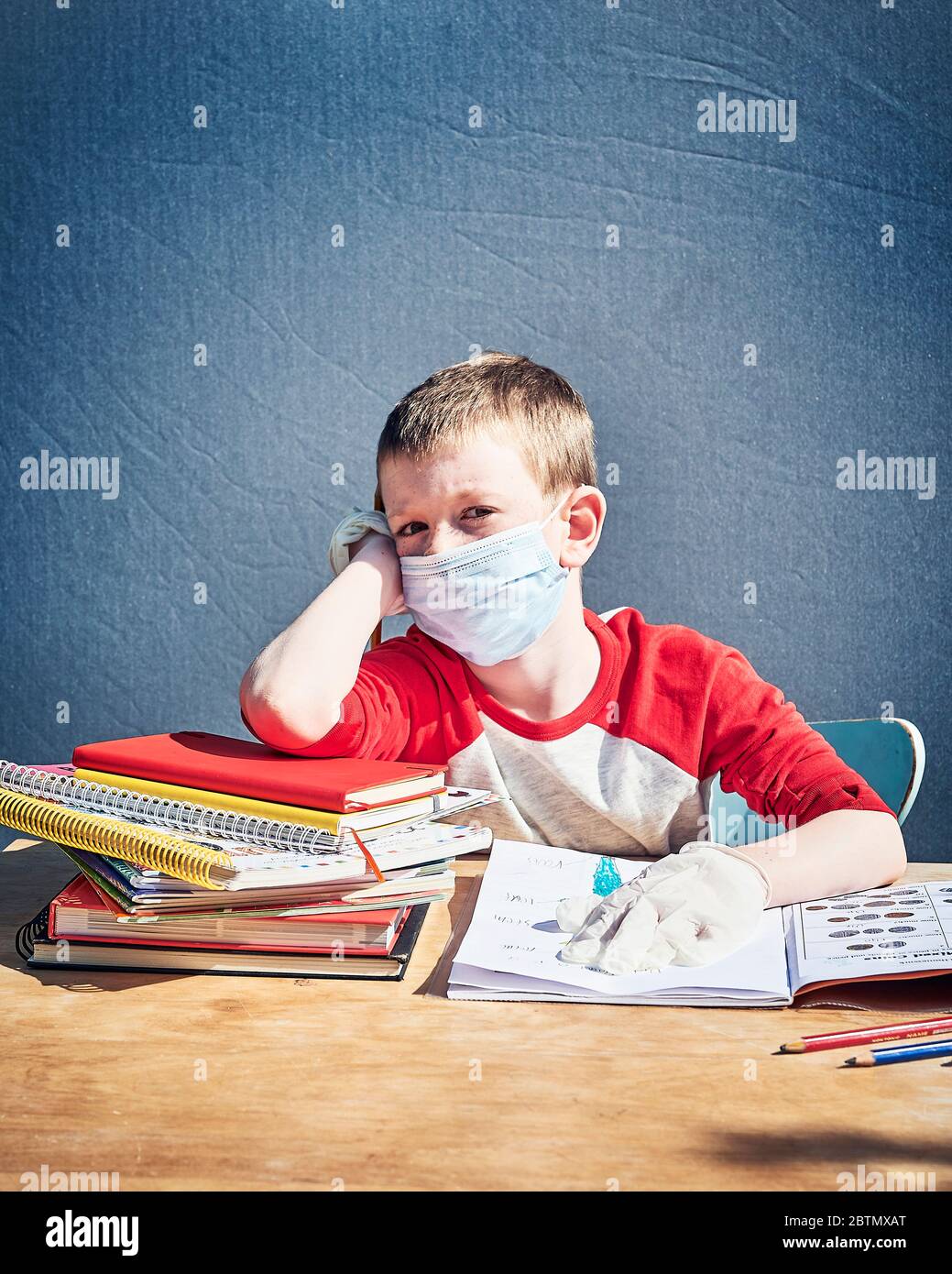 Un garçon de cinq ans est assis à un bureau d'école avec des livres portant un masque et des gants (EPI) Banque D'Images