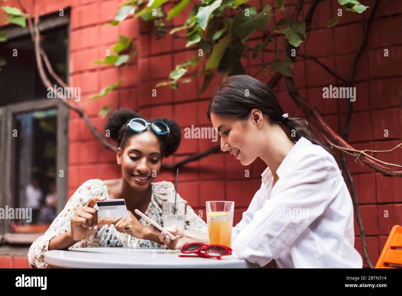 Deux jolies filles souriantes utilisant un téléphone portable et une carte de crédit tout en passant du temps ensemble dans la cour confortable du café Banque D'Images