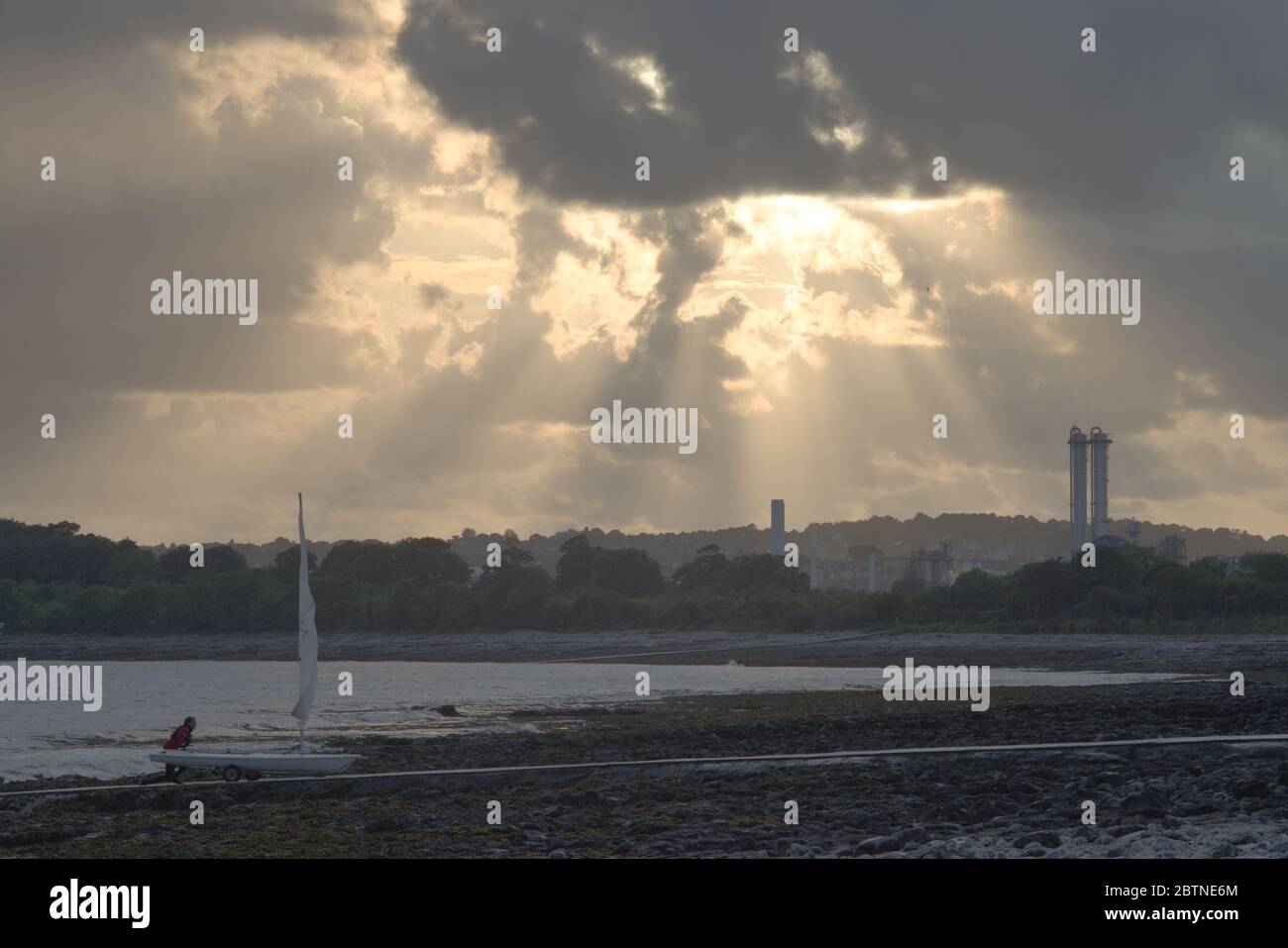 Un homme pousse son yacht sur une jetée sur une plage de sable, avec des tours industrielles d'usine derrière lui dans le sud du pays de galles Banque D'Images