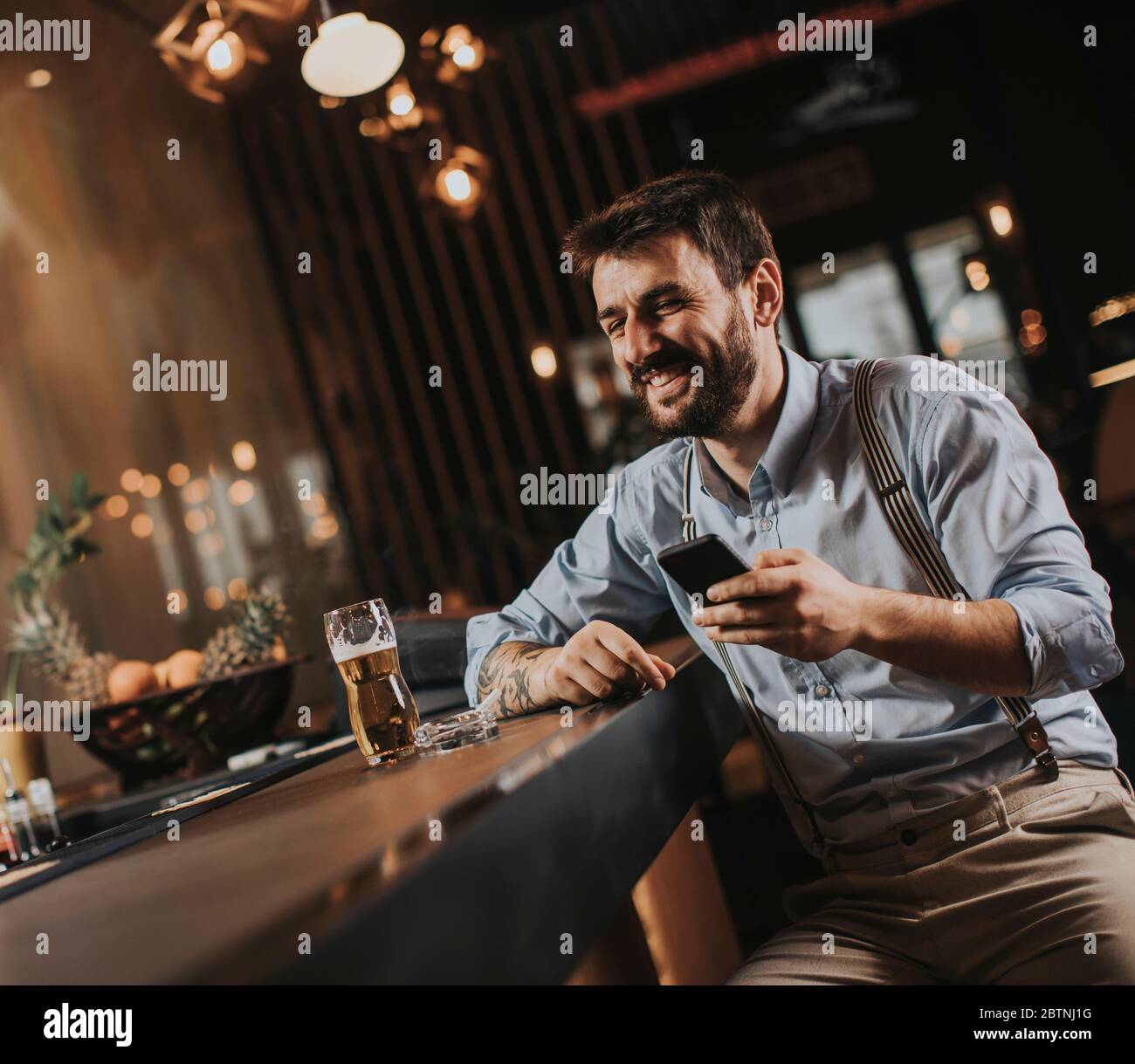 Un jeune homme heureux qui boit de la bière, fume de la cigarette et utilise le téléphone portable au pub Banque D'Images