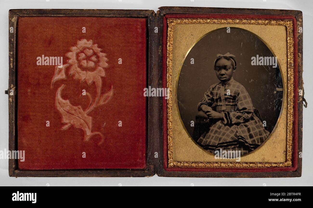 Tintype d'une fille dans une robe à carreaux. Tintype de jeune fille assise dans une robe à carreaux fantaisie avec un noeud blanc dans ses cheveux. Elle est penchée à droite et repose sur le bras d'une chaise. Le tintype est encadré dans un tapis ovale avec un cadre doré. Le tintype est dans un cas avec un motif floral sur le devant et le dos. Banque D'Images