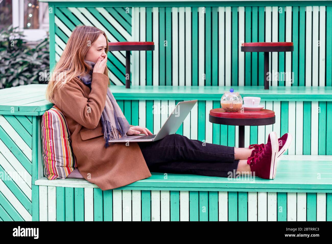 Femme caucasienne attrayante assise seule dans un café extérieur avec un fond vert vif. Une fille endormie et dépassée dans un café travaillant sur un ordinateur portable tout en déjeunant et en se réveilant Banque D'Images