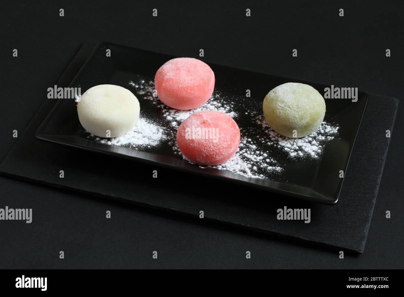 Quatre types de mochi japonais dessert - grenade au miel, thé vert matcha, fraise, noix de coco sur une assiette noire sur une table noire. Banque D'Images