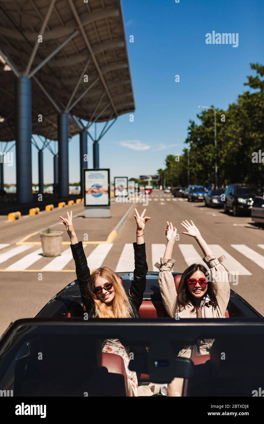 Deux filles joyeuses en lunettes de soleil se réjouissent de regarder dans l'appareil photo tout en conduisant la voiture cabriolet sur les rues de la ville en plein air Banque D'Images