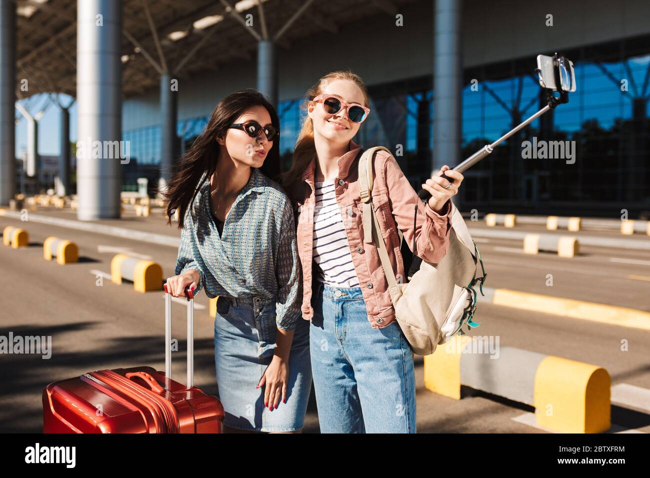 Deux jolies filles en lunettes de soleil prennent joyeusement la photo sur  un téléphone portable avec une valise et un sac à dos sur l'épaule près de  l'aéroport Photo Stock - Alamy
