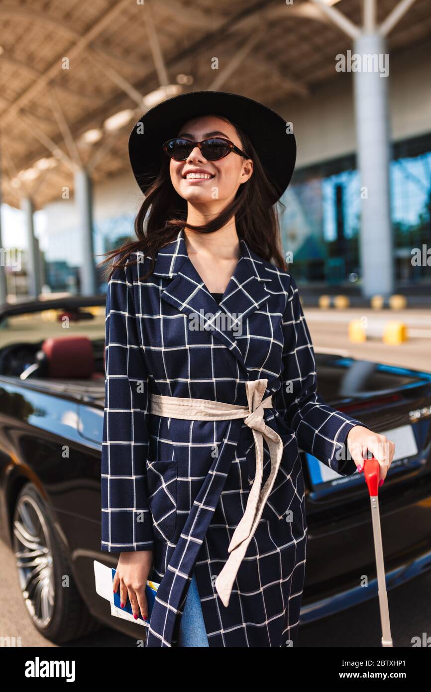 Belle fille souriante en lunettes de soleil et chapeau noir regardant avec  plaisir de côté près de l'aéroport avec cabriolet sur fond Photo Stock -  Alamy