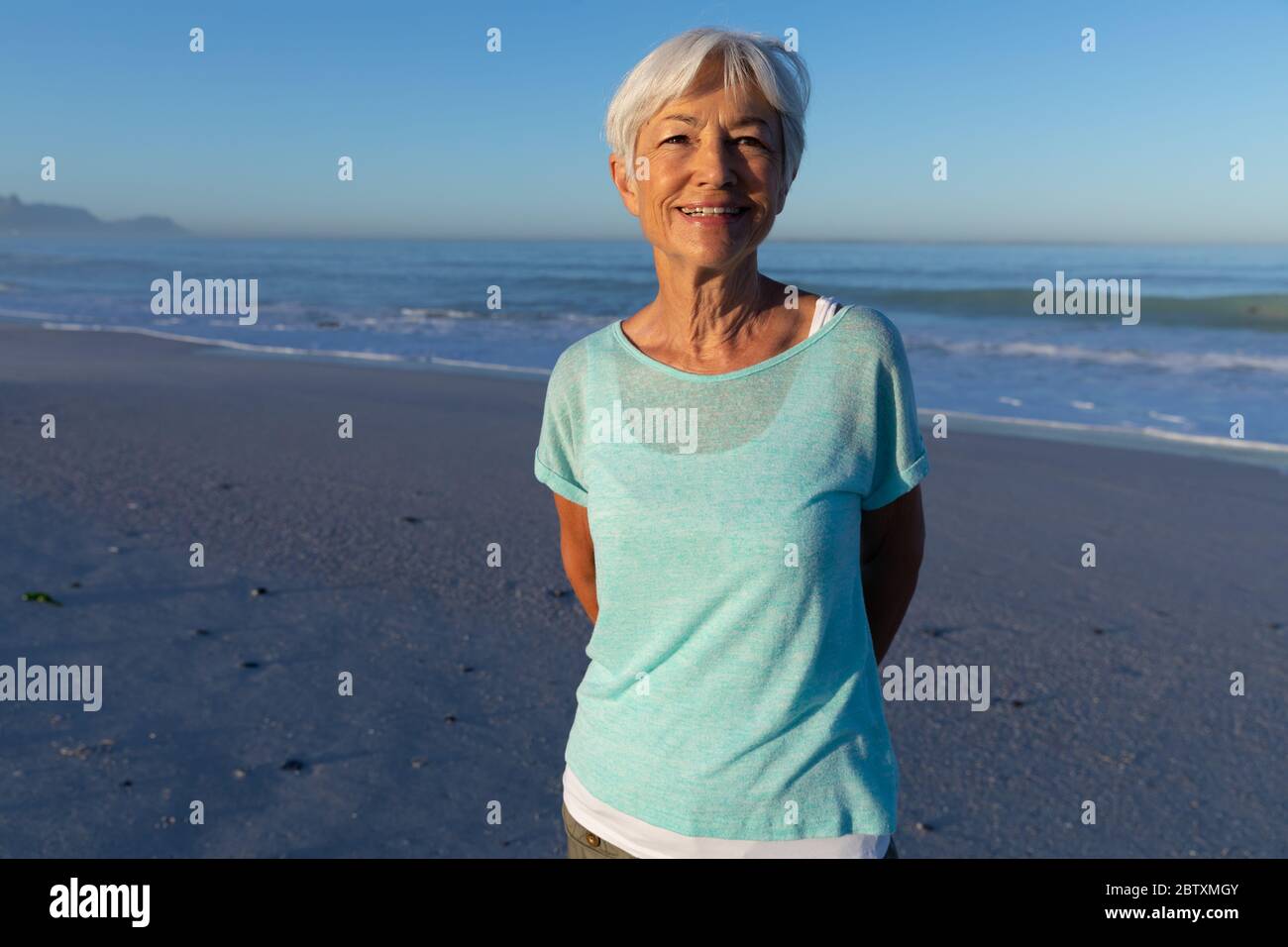 Femme caucasienne senior appréciant le temps à la plage par une journée ensoleillée, regardant la caméra et souriant avec la mer en arrière-plan Banque D'Images