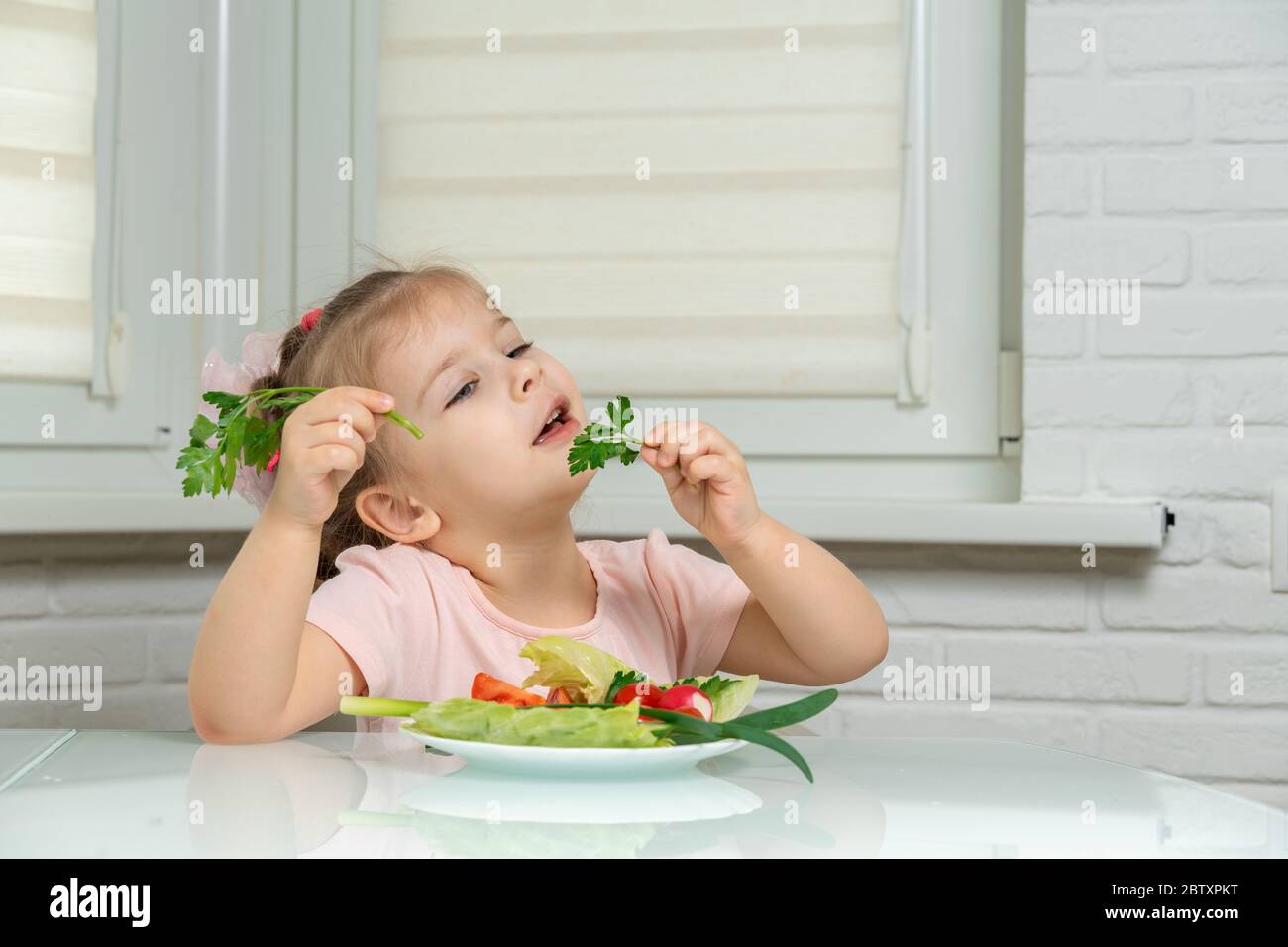 fille de 4 ans assise dans la cuisine à la table et manger des légumes, dans les mains d'une branche de persil. règles de nutrition préscolaire Banque D'Images