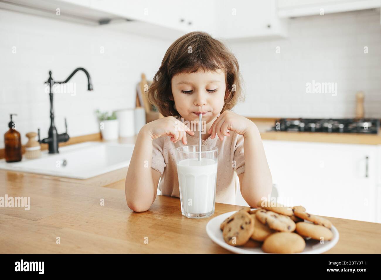 Une petite fille mignonne boit du lait avec de la paille d'acier en verre  et mange des biscuits assis à la table de la cuisine. Réduire l'utilisation  de plastique chez les enfants